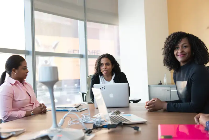 Three colleagues discussing work around a meeting table with laptops